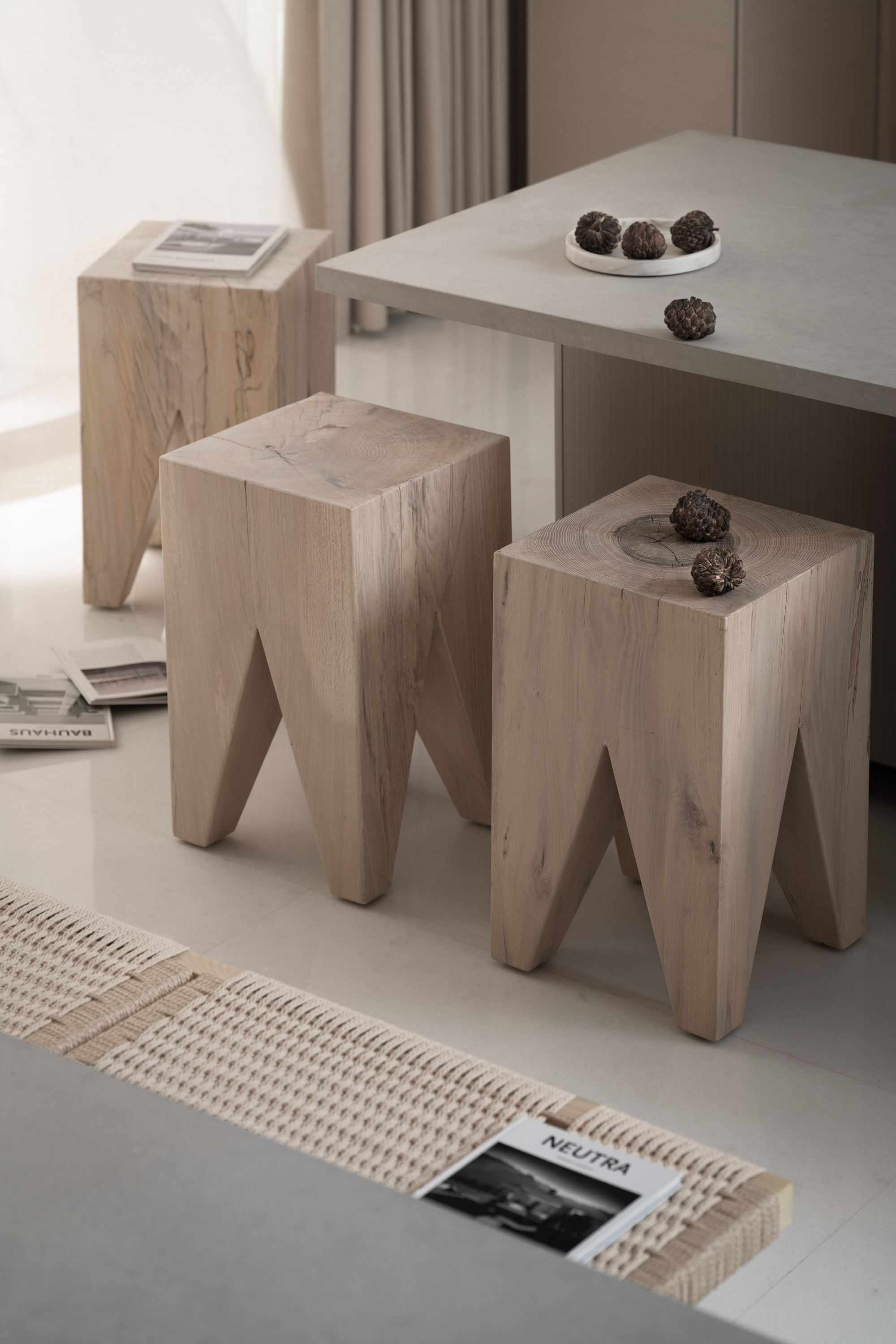 Close-up of pale wooden tripod stools tucked beneath a dining table with ceramic objects placed on top